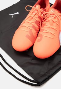 Bottines de soccer orange vif avec un upper texturé et des accents noirs, posées sur un sac à cordon noir avec un logo blanc.
