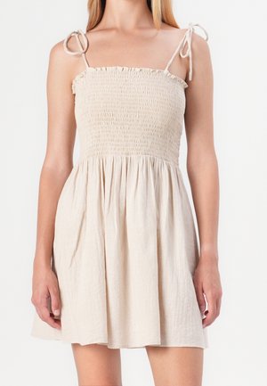 Mujer con vestido beige sin mangas, con corpiño fruncido y tirantes anudados en los hombros, posando frente a un fondo blanco liso.