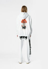 Sudadera blanca de gran tamaño con un gráfico de un perro de dibujos animados colorido en la parte posterior, combinada con pantalones de chándal blancos que presentan letras y acentos en negro.