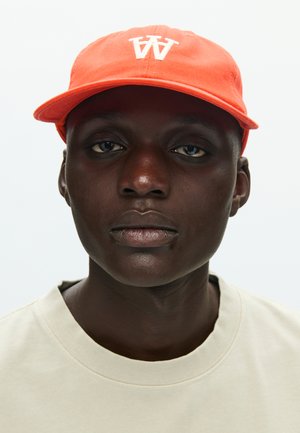 Orange cotton cap with white "W" logo, paired with a light beige crew-neck t-shirt. Close-up of the head and shoulders.