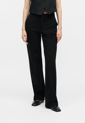 Osoba nosiaca čierne na mieru šité široké nohavice, čiernu zapínanú vestu a čierne topánky so štvorcovou špičkou na jednoduchom pozadí.