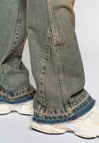 Jean bleu délavé à jambes larges avec ourlets effilochés, porté au-dessus de baskets blanches à semelles épaisses et détails en mesh.