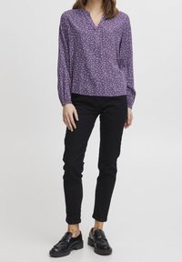 Lila blus med vita prickar, v-ringning och långa ärmar som samlas vid handledarna, kombinerad med svarta slim-fit byxor och svarta loafers.