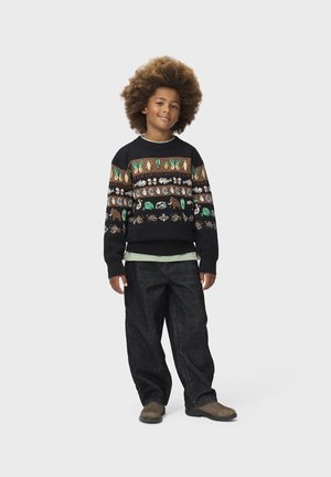 Kind mit großem lockigem Haar steht, trägt einen gemusterten Pullover mit Tieren und Pflanzen, schwarze Hose und braune Schuhe vor schlichtem Hintergrund.