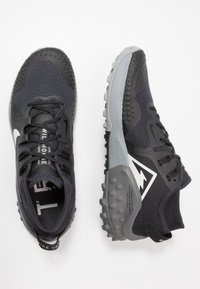 Zwarte en grijze Nike trail running schoenen met gestructureerde zolen, vetersluiting en wit driehoekig logo aan de zijkant, getoond vanuit boven- en zijaanzicht.