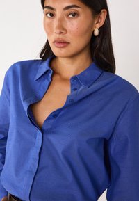 Camicia azzurra con bottoni e colletto, realizzata in tessuto strutturato e con sei bottoni sulla parte anteriore. Il modello ha capelli scuri e indossa orecchini.