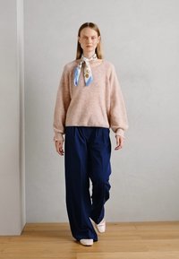 Lichtroze, getextureerde oversized trui met losse mouwen, gecombineerd met marineblauwe wijdvallende broek, en een gestreepte zijden sjaal die om de hals is geknoopt.