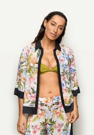 Kobieta w koszuli i spodniach w kwiatowy wzór z czarnym obszyciem, oliwkowo-zielonym topem bikini, stojąca na jednolitym jasnym tle.