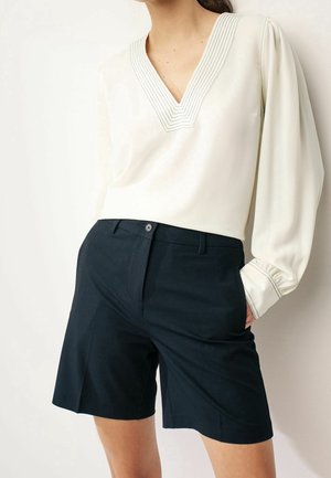 Femme portant un chemisier blanc à manches longues avec un col en V cousu et un short noir sur mesure jusqu'aux genoux, debout avec la main dans la poche.