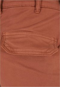 Gros plan d’un tissu couleur rouille avec une patte de poche cousue, un passant de ceinture et des détails de couture visibles sur un vêtement.