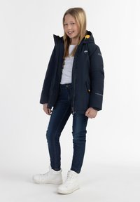 Marinblå isolerad jacka med en gul huva, utrustad med en framdragkedja och sidofickor. Kombination med blå jeans och vita sneakers.