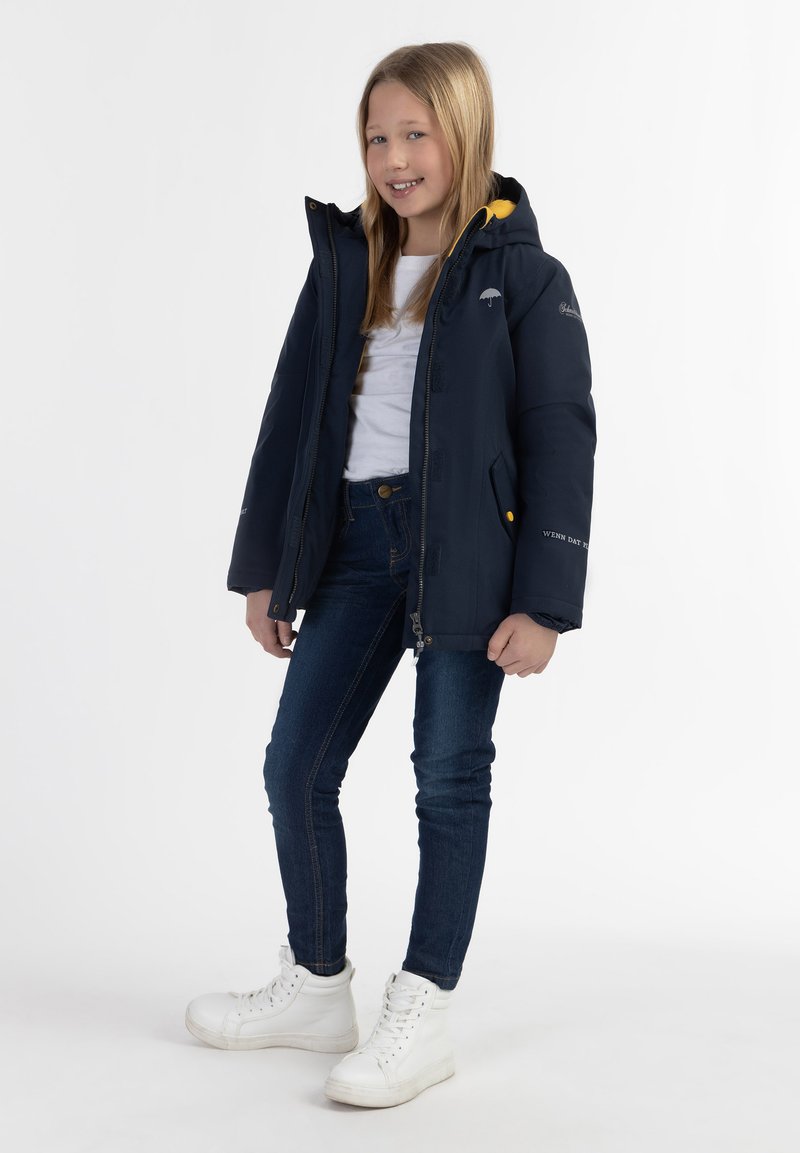 Marinblå isolerad jacka med en gul huva, utrustad med en framdragkedja och sidofickor. Kombination med blå jeans och vita sneakers.