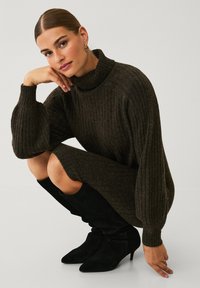 Dunkelgrünes, geripptes Strickkleid mit hohem Kragen und übergroßen Ärmeln, kombiniert mit kniehohen, schwarzen, samtigen Stiefeln mit spitzem Absatz.