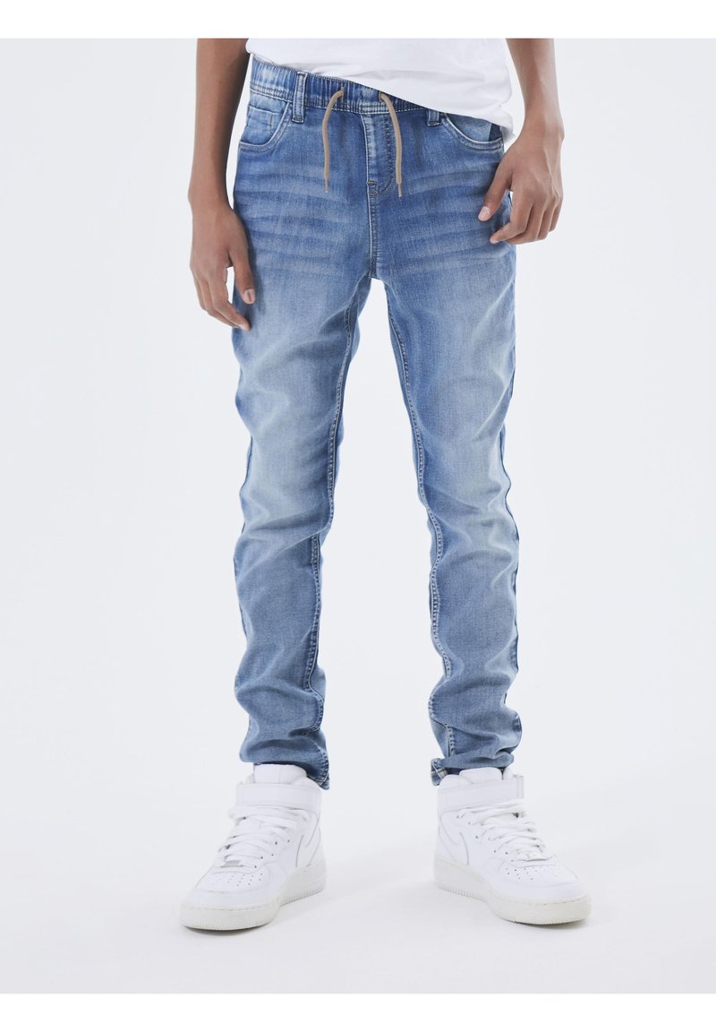 Personne portant un jean jogger en denim bleu clair avec une taille élastique et un cordon marron, associé à des baskets montantes blanches.