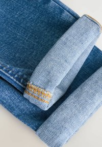 Denim hlače z zavihanimi robovi, v srednje modri barvi, z diskretno teksturo in kontrastnim rumenim šivom ob robu.