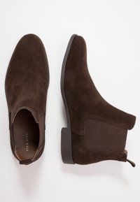Botines de ante marrón con paneles laterales elásticos, punta redondeada y suela plana de goma. Diseño clásico adecuado para el uso diario.