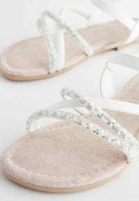 Sandali bianchi con una fascia gioiello, che presenta decorazioni trasparenti e simili a perle. Base in camoscio rosa chiaro e design piatto.