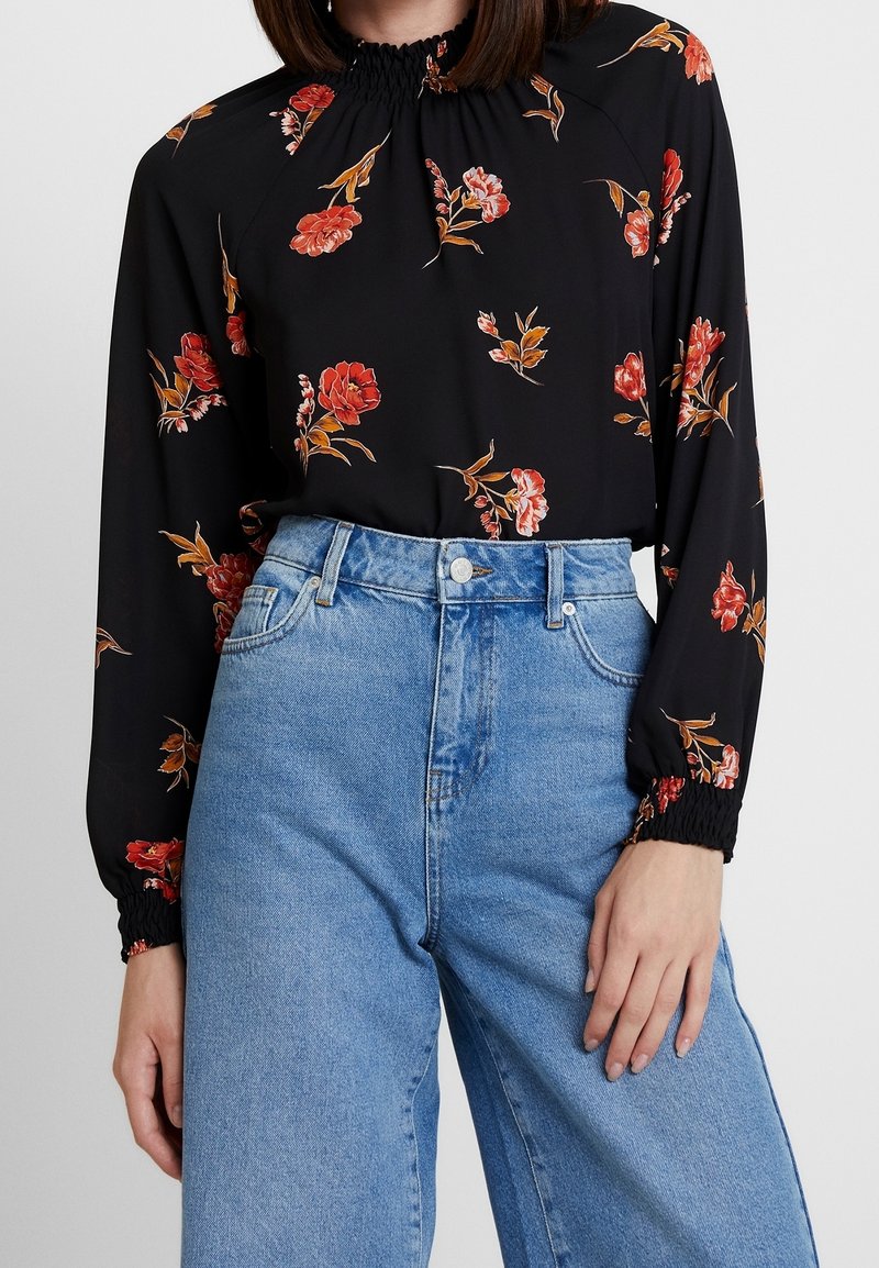 Donna che indossa una blusa nera a maniche lunghe con un motivo floreale rosso e con polsini elasticizzati, infilata in jeans chiari a vita alta.