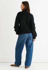 Donna in piedi di lato, rivolta a sinistra, che indossa un maglione nero a maglia grossa, jeans blu larghi e scarpe basse con stampa leopardata.