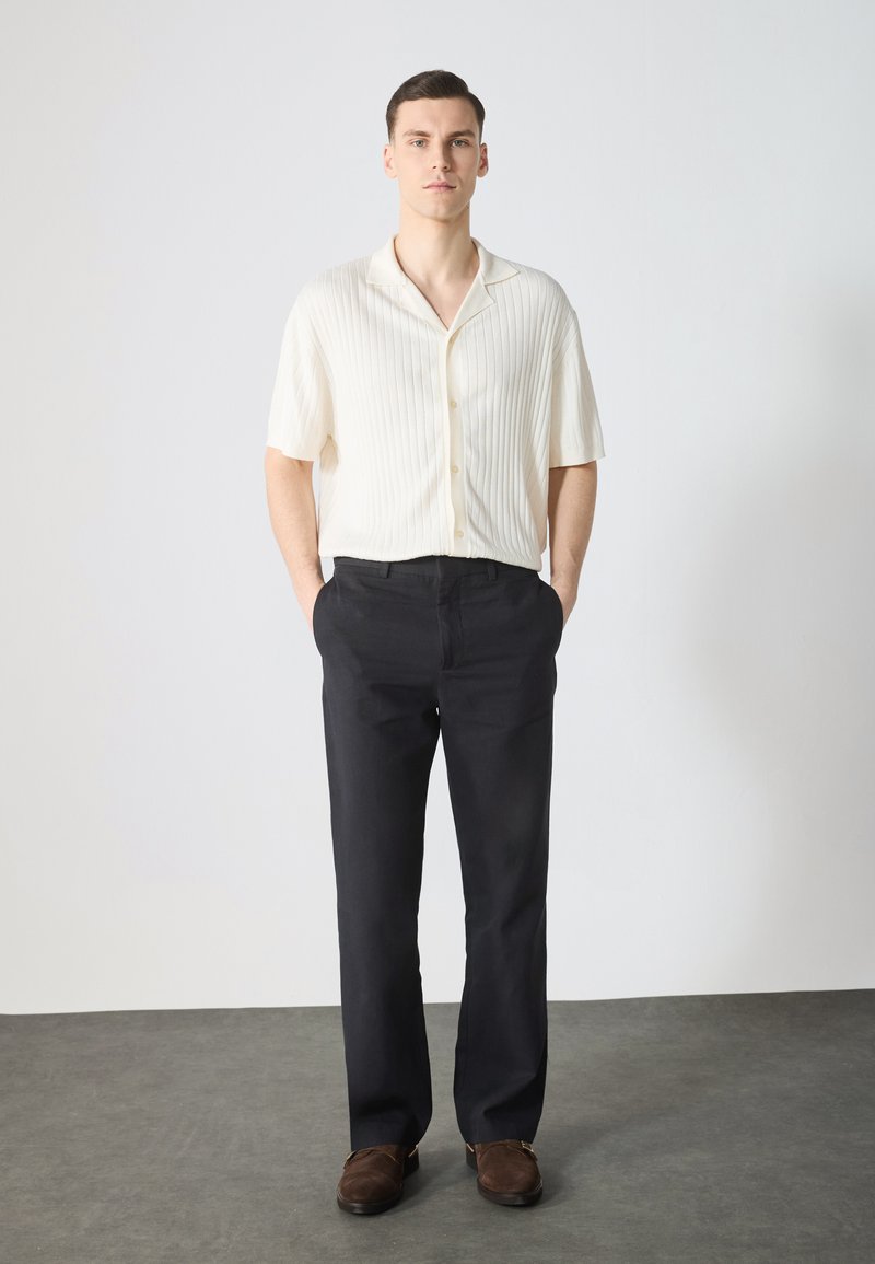 Chemise à manches courtes en crème à côtes avec des boutons, associée à un pantalon sombre et des chaussures à enfiler brunes. Arrière-plan uni avec une texture minimale.