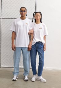 Two individuals wear matching white T-shirts featuring a colorful graphic on the left chest. They pair with light blue and dark denim jeans.
