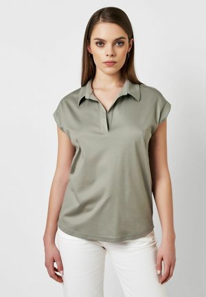 Kurzarm-Poloshirt in gedämpftem Grün, aus glattem Stoff gefertigt. Verfügt über eine knopffreie Blende und einen dezenten Glanz. Kombiniert mit weißen Hosen.