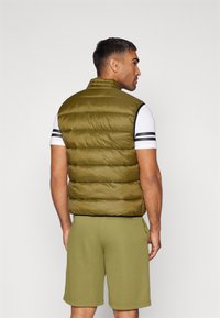 Olivgrüne Steppweste mit vertikaler Naht, hohem Kragen und Reißverschlüssen, kombiniert mit hellgrauen Shorts und einem weißen T-Shirt mit schwarzen Streifen.
