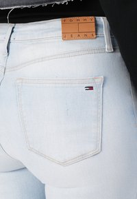 Svetlomodré denimové džiny s priliehavým strihom, vybavené koženou nášivkou s označením "TOMMY JEANS" a malým vyšívaným logom v červenej, bielej a modrej farbe.