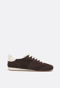 Sneaker a profilo basso in suede marrone scuro con lacci bianchi, toppa sul tallone bianca sporca e suola in gomma marrone chiaro, mostrato dal lato esterno.