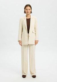 Beige oversized jakkesæt med en enkeltstående blazer og vide bukser, parret med en tætsiddende brun top og vævede brune sko.