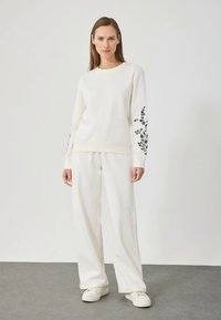 Valge puuvillane pusa musta lillelise tikandiga varrukaid, sobitatud lahtiste valgete pükstega ja valgete tossudega.