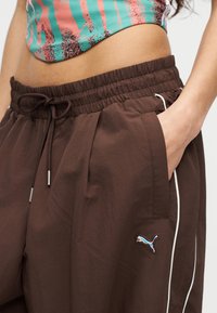 Κοντινό πλάνο γυναίκας που φορά καφέ αθλητικό παντελόνι Puma με κορδόνι στη μέση και το χέρι στην τσέπη, συνδυασμένο με ένα πολύχρωμο κοντό τοπ.