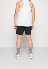 Camiseta blanca sin mangas, pantalones cortos negros, calcetines grises con textura y zapatillas beige, vistos desde atrás sobre un fondo neutro.