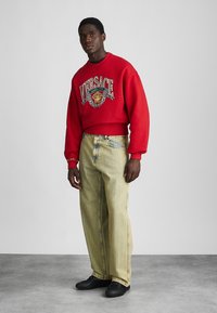 Sweatshirt rouge avec le logo "Versace" et un graphique de lion, associé à un jean en denim clair. Des chaussures noires complètent le look.