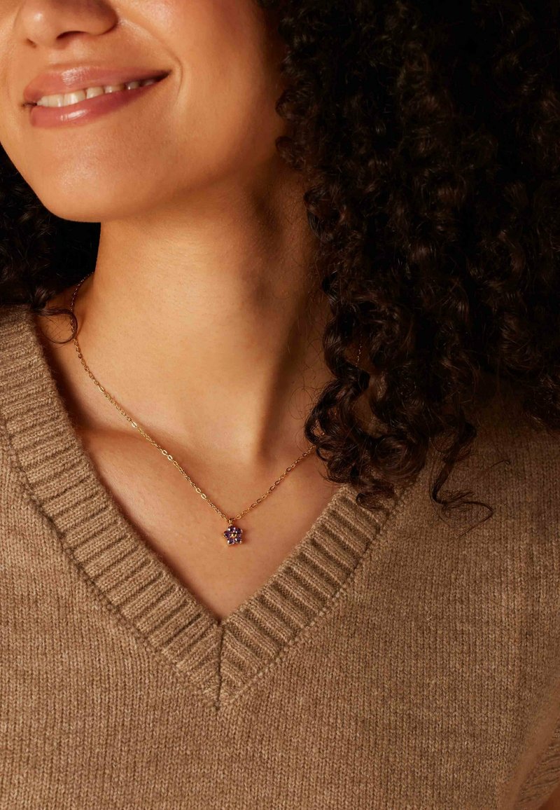 Gouden ketting met een bloemenvormige hanger met paarse accenten, gedragen over een beige V-halstruijka met ribbelstructuur.