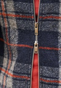 Wollmischung-Jacke mit navy- und grau-karierter Musterung, die orange Linien aufweist. Zentrale Reißverschluss mit metallischen Details, über einem roten Oberteil getragen.