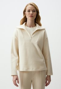 Helles Creme-Pullover aus strukturiertem Stoff, mit hohem Kragen, Reißverschluss und lockerer Passform. Kombiniert mit beigen, enganliegenden Hosen.