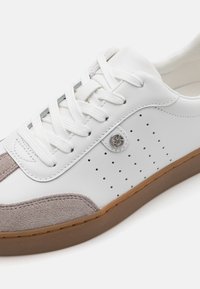 Sneaker bianco con punta in suede taupe, lati perforati e lacci tono su tono. Presenta un accento in metallo con logo e una suola in gomma marrone.