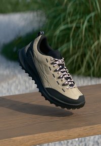 Beżowe i czarne buty trekkingowe z różowymi sznurówkami uniesione nad drewnianą powierzchnią, z zielonymi roślinami i żwirem w tle.