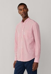 Chemise boutonnée à rayures rouges et blanches, manches longues, col ajusté, poche poitrine et ourlet arrondi. Portée avec un pantalon bleu marine.
