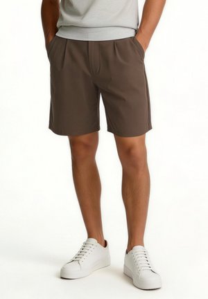 Mann trägt braune Shorts, weiße Sneakers und ein hellgraues T-Shirt, steht mit den Händen in den Taschen vor weißem Hintergrund.