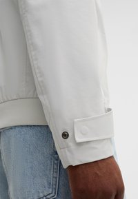 Weiße leichte Jacke mit Reißverschluss, gerippten Bündchen und Druckknopfdetail am Ärmel. Glatte Textur mit minimalistischer Gestaltung.