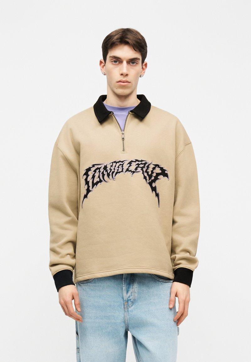 Νέος άνδρας που φοράει μπεζ πουλόβερ με φερμουάρ ως το λαιμό, με μαύρο γιακά και μανσέτες, ανοιχτό μπλε τζιν, στέκεται μπροστά σε απλό λευκό φόντο.