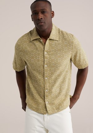 Férfi, rövid ujjú, gombos ingben, mustár és fehér geometriai virágmintával, fehér nadrággal, egyszerű háttér előtt állva.