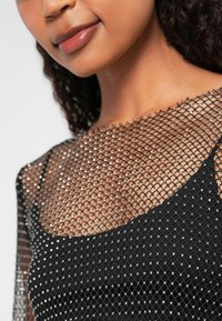 Svart plagg med et gjennomsiktig, glitrende mesh-overlegg som har et diamantmønster, som fremhever et glatt, tettsittende underlag.
