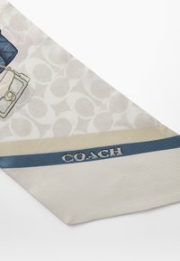 Hedvábný šátek v jemné krémové barvě s vzorovaným pozadím, doplněný modrým akcentním pruhem a modrým nápisem "COACH".