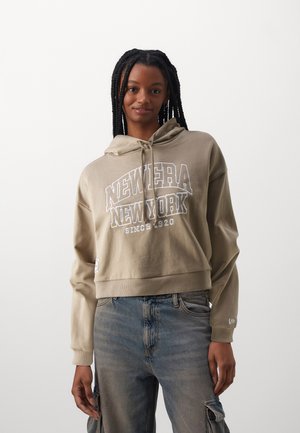 Sudadera cropped beige con "NEWERA NEW YORK" impreso en blanco, que presenta una capucha con cordón y puños acanalados, combinada con jeans de lavado claro.