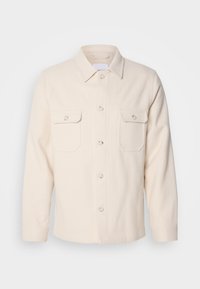 Light beige button-up shirt made of textured fabric; features two chest pockets, a pointed collar, and long sleeves with buttoned cuffs.