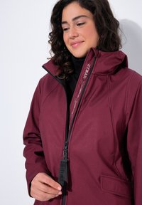 Chaqueta impermeable burdeos con cuello alto, cierre de cremallera y tela texturizada. Presenta tirador de cremallera con marca y bolsillos laterales.