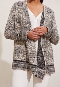 Stickad cardigan med intrikata svarta och krämfärgade mönster, öppen framsida och kontrasterande mönstrade ärmslut och fåll. Mjuk textur, avslappnad passform.